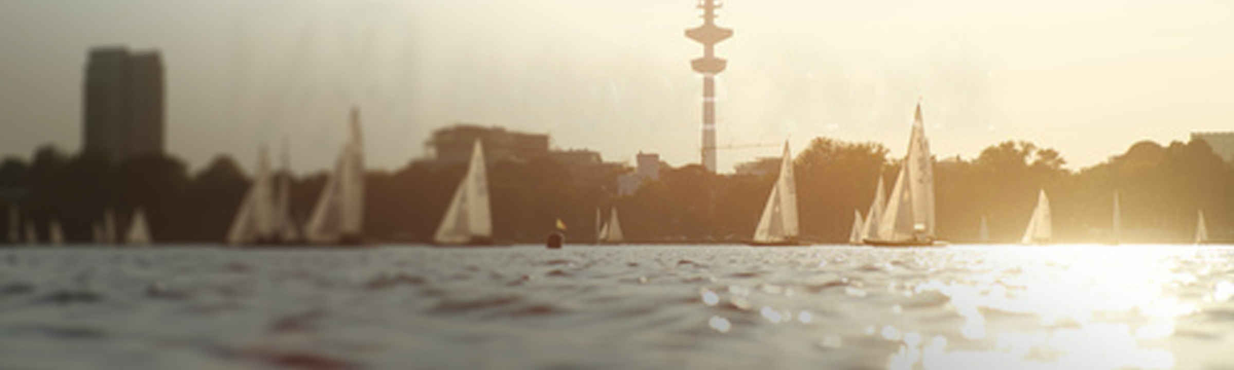 Segelboote auf der Binnenalster in Hamburg bei Sonnenuntergang, Fernsehturm im Hintergrund, ruhige Wasseroberfläche.