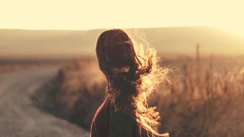 Frau steht mit Wind in den Haaren auf Feld bei Sonnenuntergang.