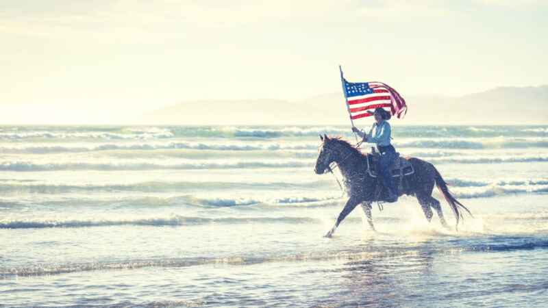 Reiter mit US-Flagge reitet auf einem Pferd entlang des Meeres, bei strahlendem Sonnenschein.