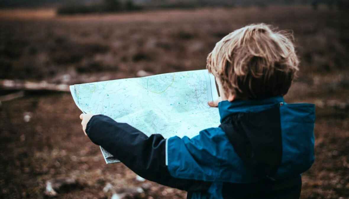 Junge mit blauer Jacke hält eine Karte im Freien, erkundet die Landschaft und sucht den Weg.