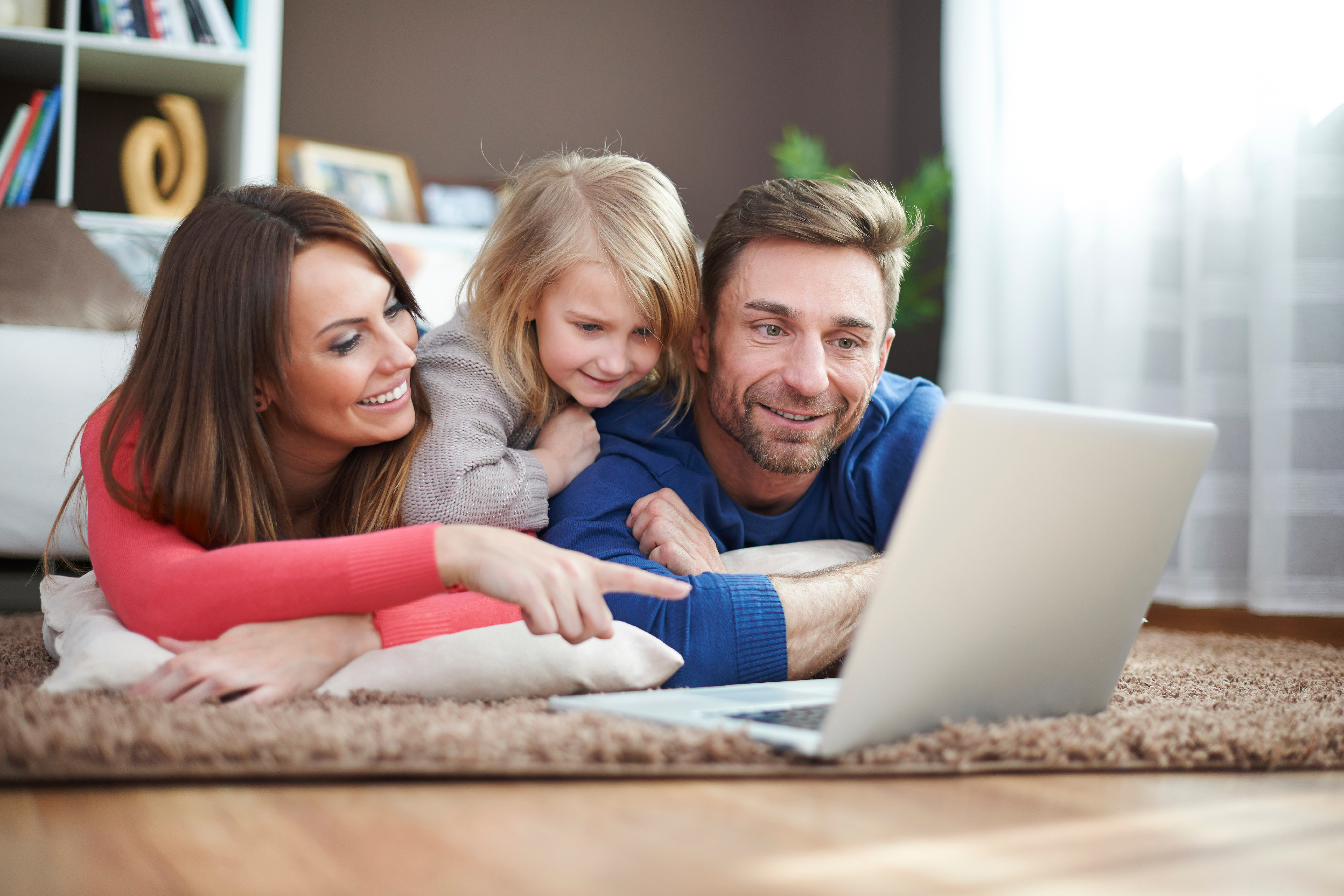 Familie liegt auf dem Teppich vor einem Laptop und lächelt zusammen.