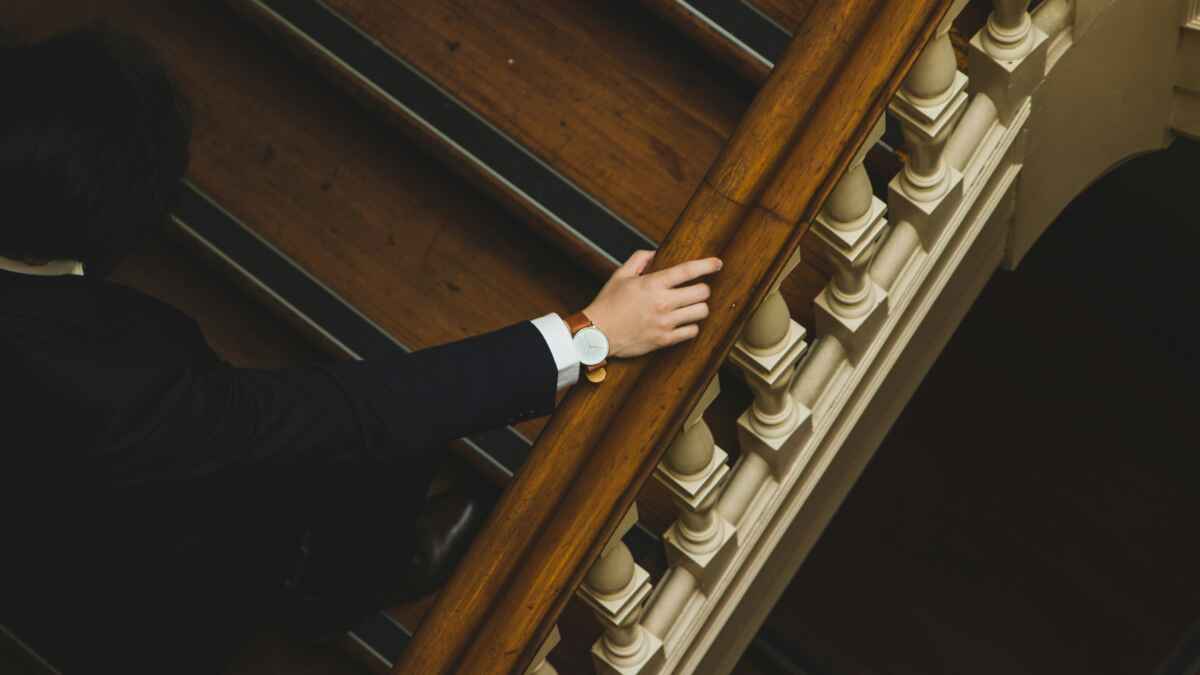 Person in dunklem Anzug steigt hölzerne Treppe hinunter, Hand am Geländer, elegante Uhr am Handgelenk.