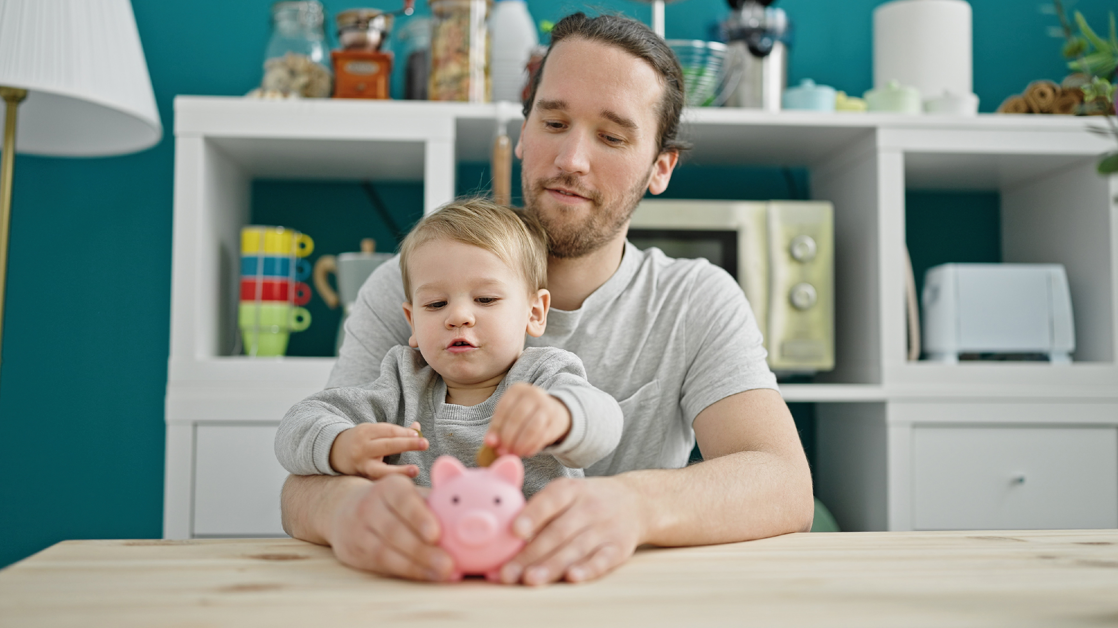 Vater und Kind am Tisch mit Sparschwein, helle Küche im Hintergrund, beide lächeln und interagieren glücklich.