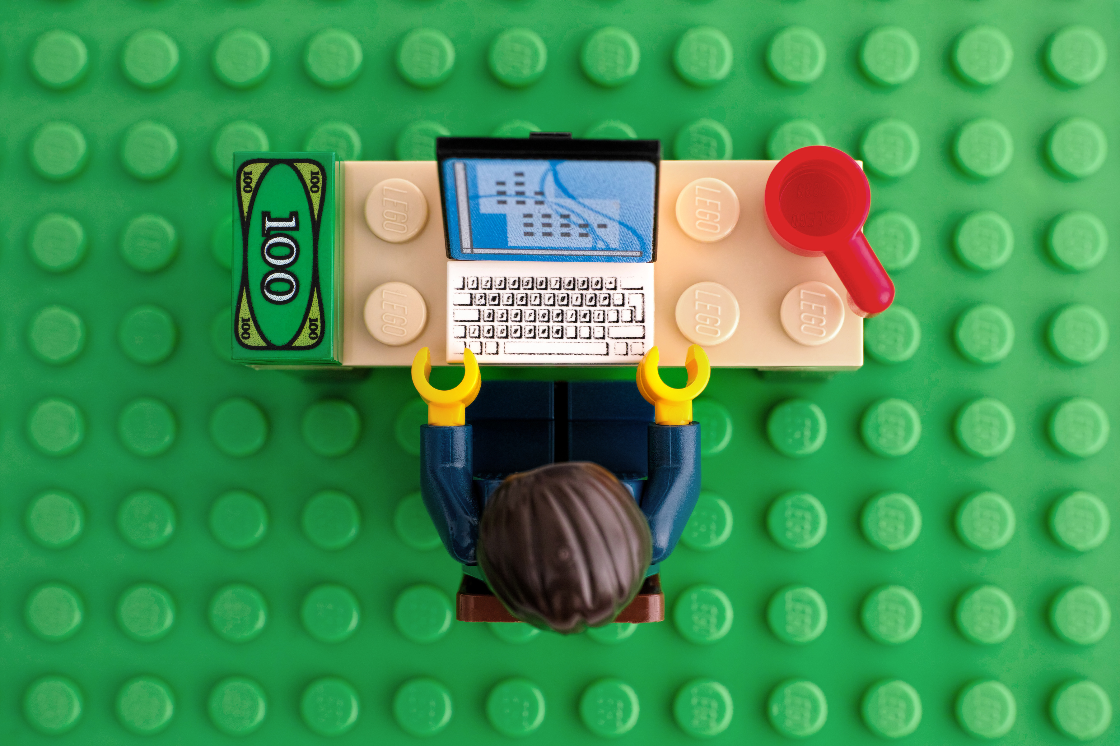 LEGO-Minifigur sitzt am Schreibtisch mit Laptop, roter Tasse und Geldschein auf grüner Basisplatte.