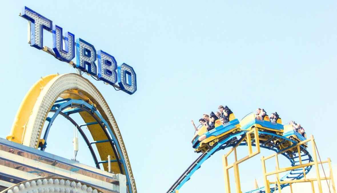 Achterbahn mit TURBO-Schild und Wagen voller fröhlicher Menschen auf einer steilen Abfahrt bei klarem Himmel.