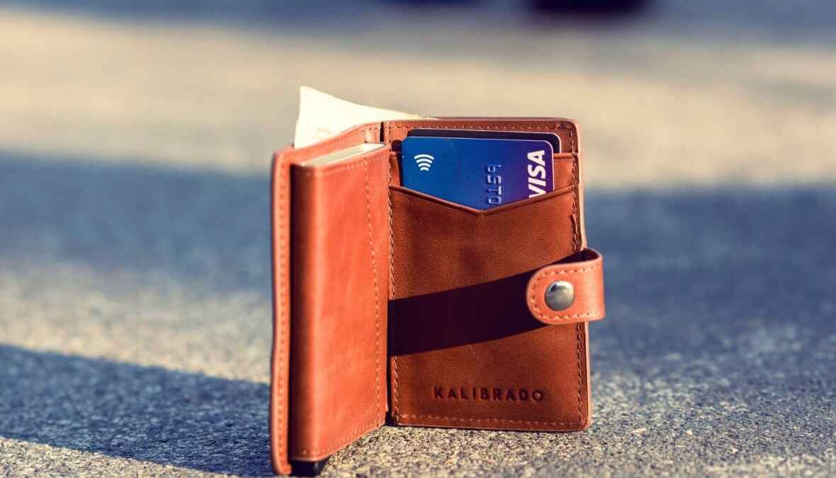 Braune Ledergeldbörse mit geöffneter Klappe, darin eine Visa-Karte und ein Geldschein, auf Asphalt liegend.