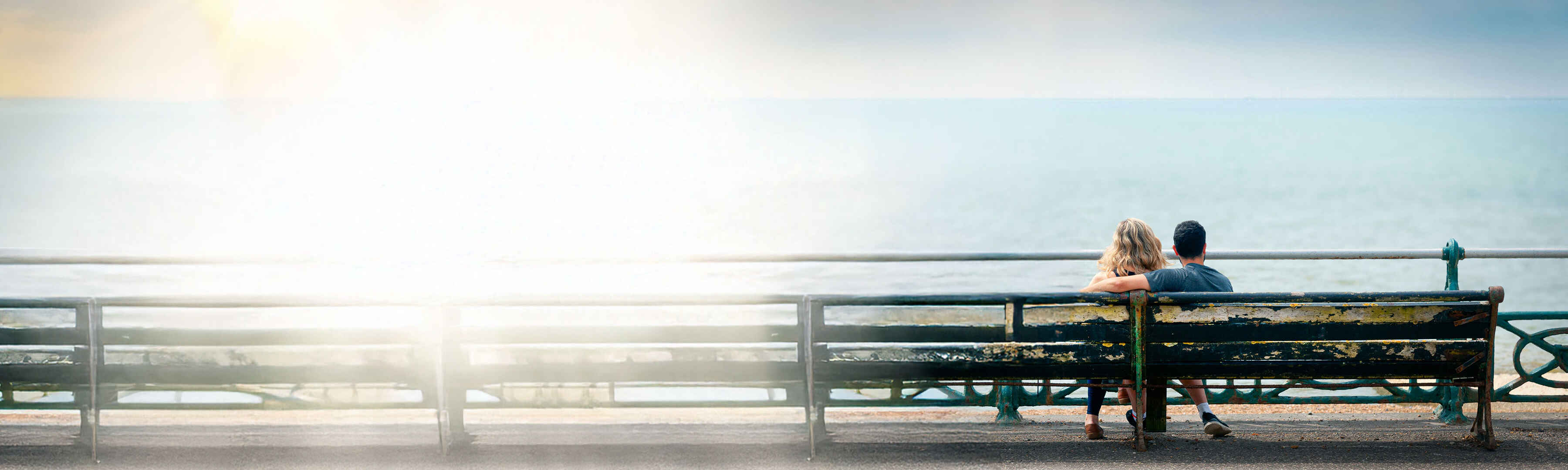 Pärchen sitzt auf einer Bank am Meer und genießt den Ausblick in der Sonne.
