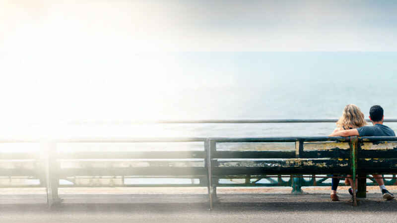 Pärchen sitzt auf einer Bank am Meer und genießt den Ausblick in der Sonne.
