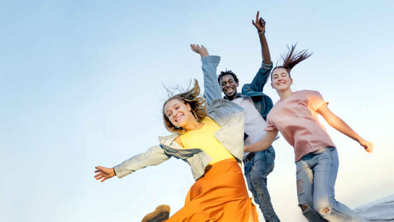 Drei fröhliche Menschen springen mit ausgestreckten Armen unter klarem Himmel. Dynamische Sommerstimmung.