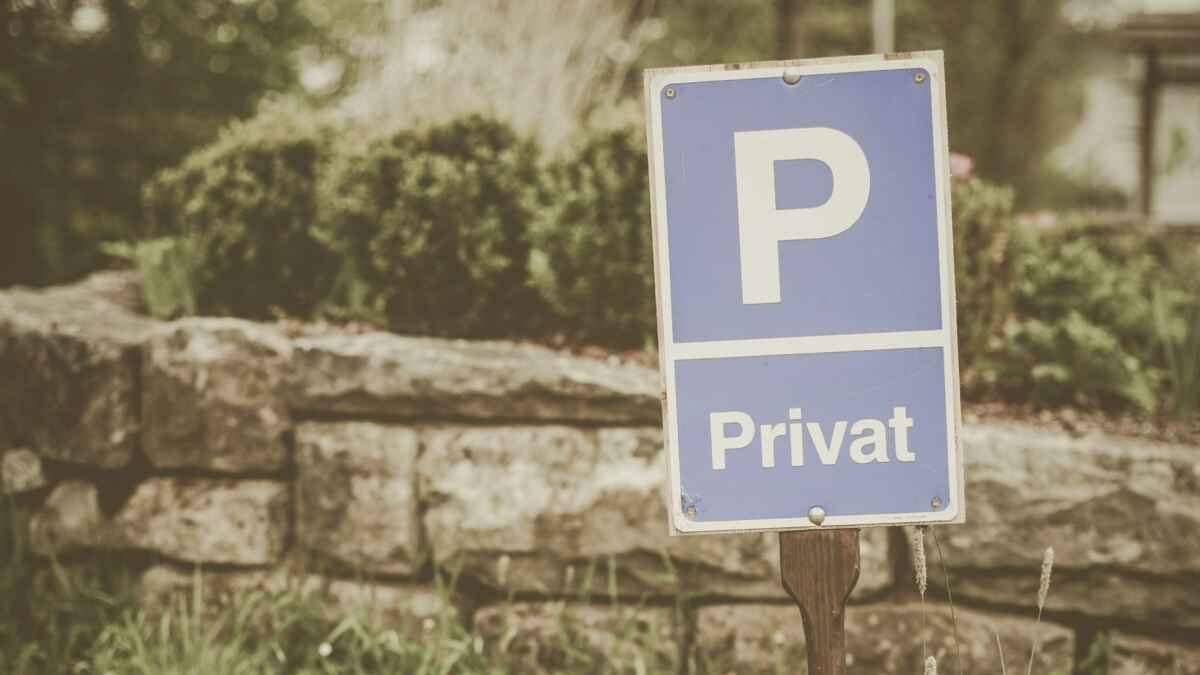Blaues Schild mit der Aufschrift P Privat auf grasbewachsenem Gelände vor einer Steinmauer.