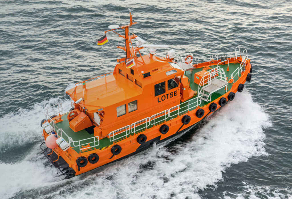 Orangefarbenes Lotsenboot mit deutscher Flagge fährt durch das Meer, hinterlässt Wellenspur im Wasser.