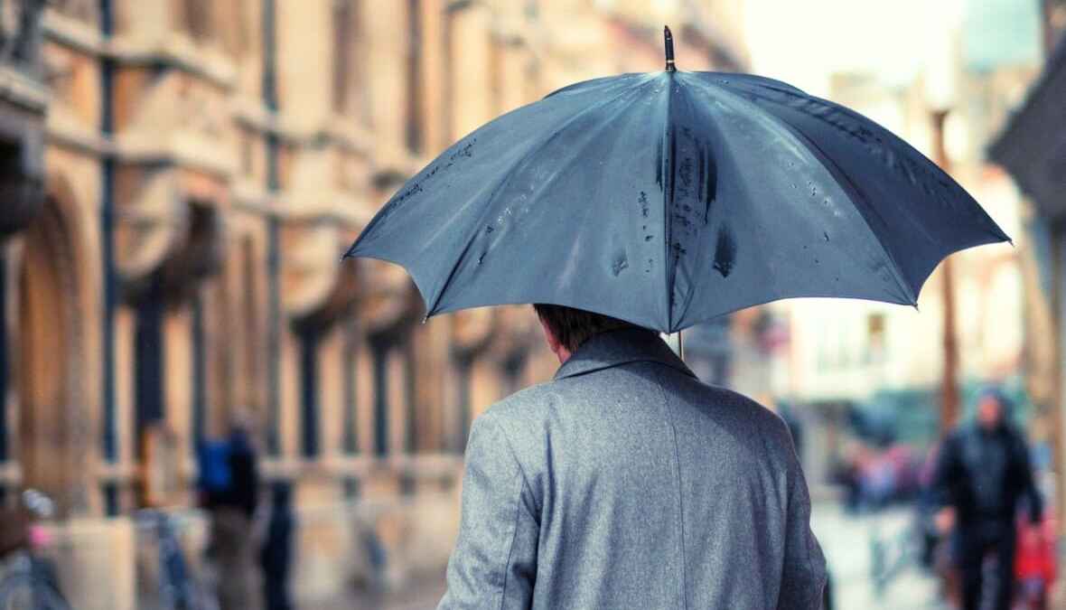Person mit grauem Mantel und schwarzem Regenschirm in städtischer Umgebung an einem regnerischen Tag.