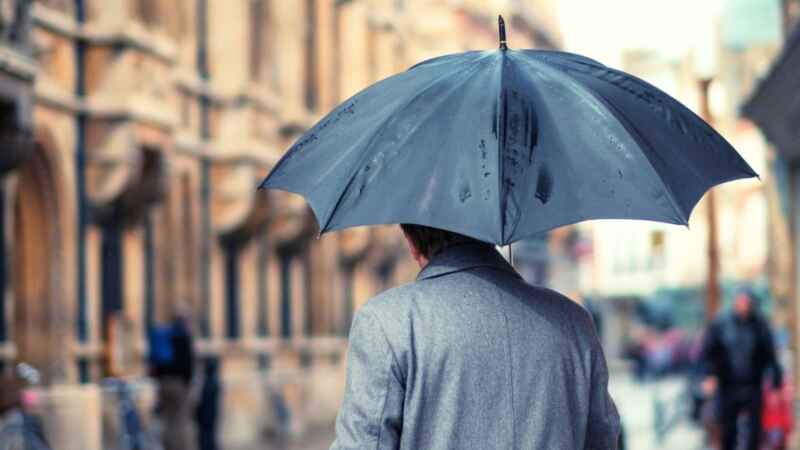 Person mit grauem Mantel und schwarzem Regenschirm in städtischer Umgebung an einem regnerischen Tag.