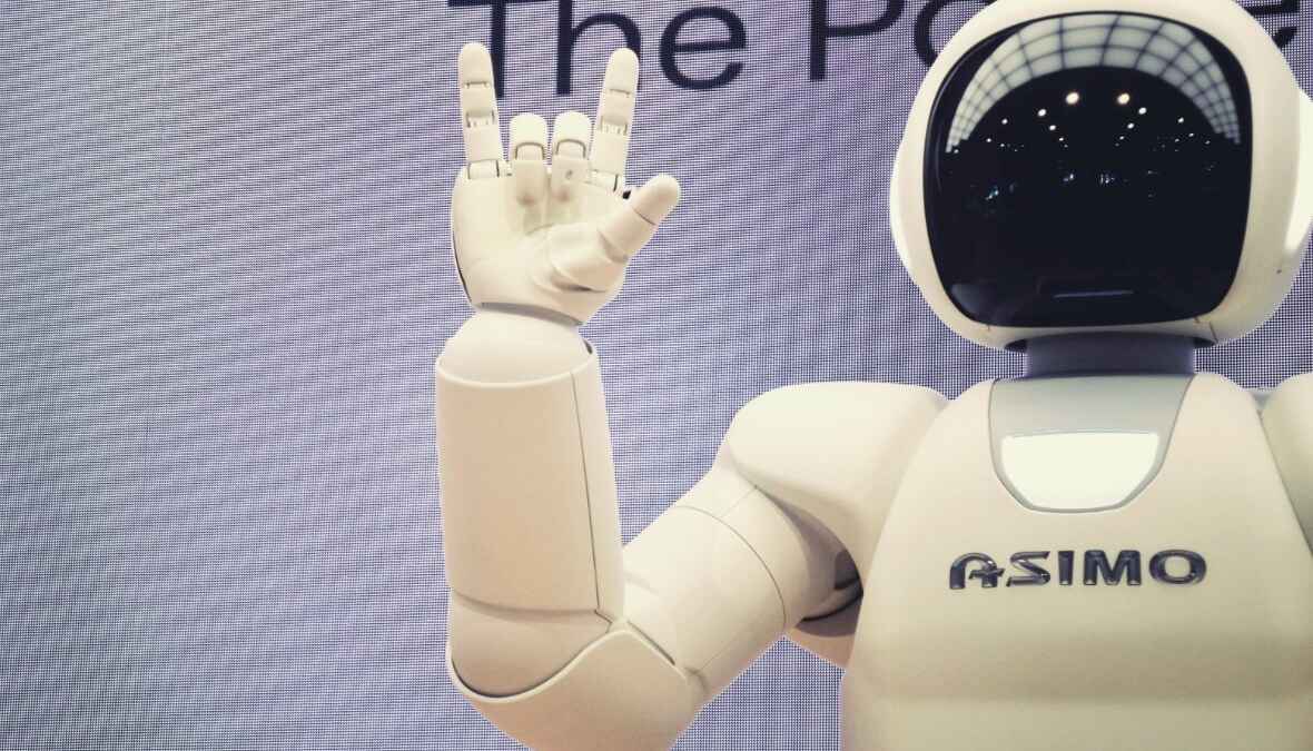 ASIMO-Roboter hebt Hand in Rock'n'Roll-Geste vor grauem Hintergrund.