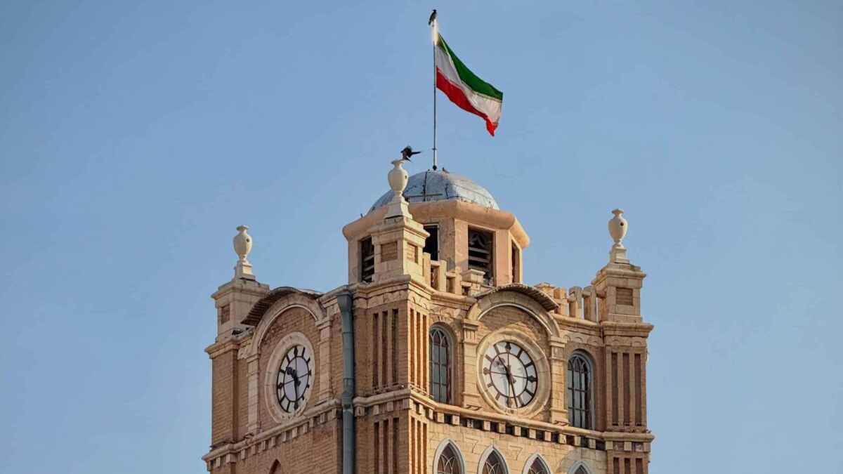 Uhrturm in Tabriz mit iranischer Flagge, blauer Himmel im Hintergrund.