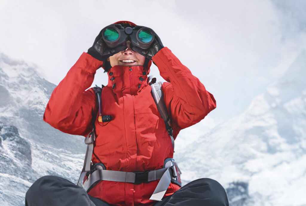 Person in roter Jacke beobachtet mit Fernglas in schneebedeckten Bergen.