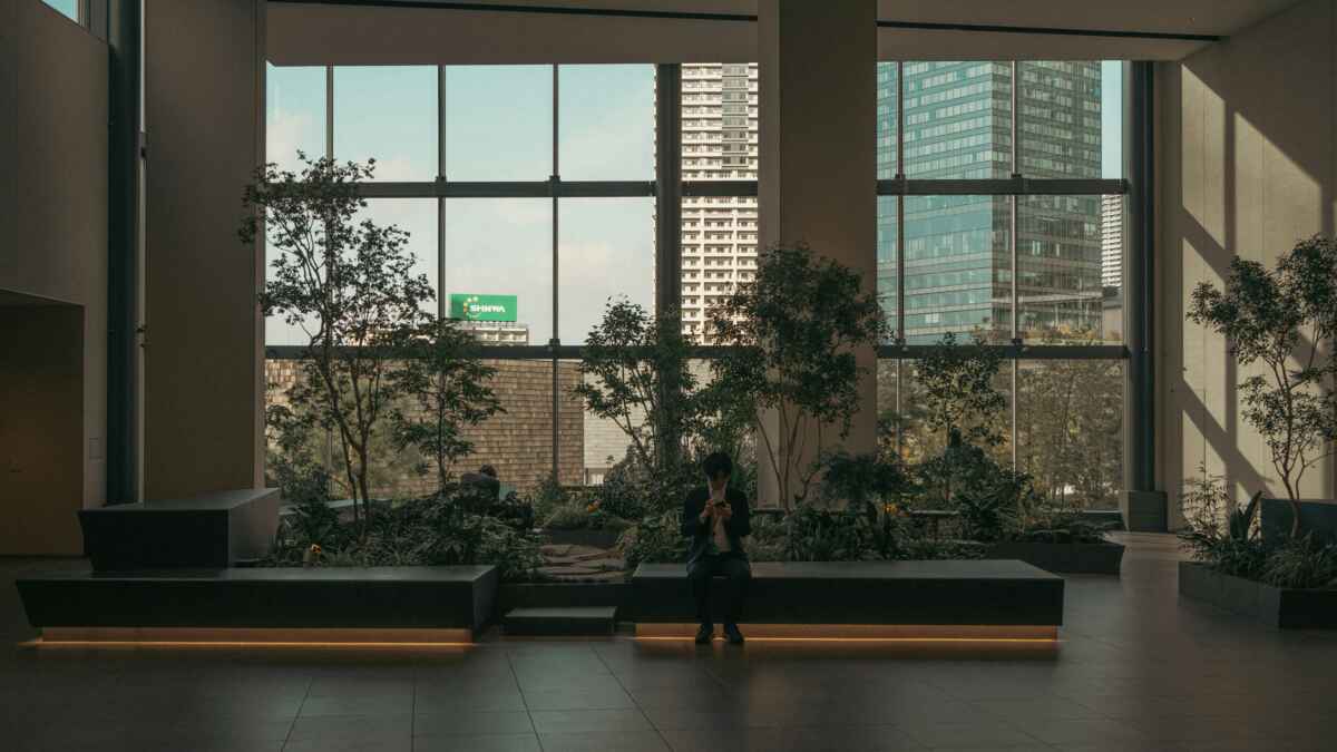 Mann in modernem Bürogebäude, sitzt auf Bank mit Smartphone, große Fenster mit Stadtblick und Pflanzen im Hintergrund.