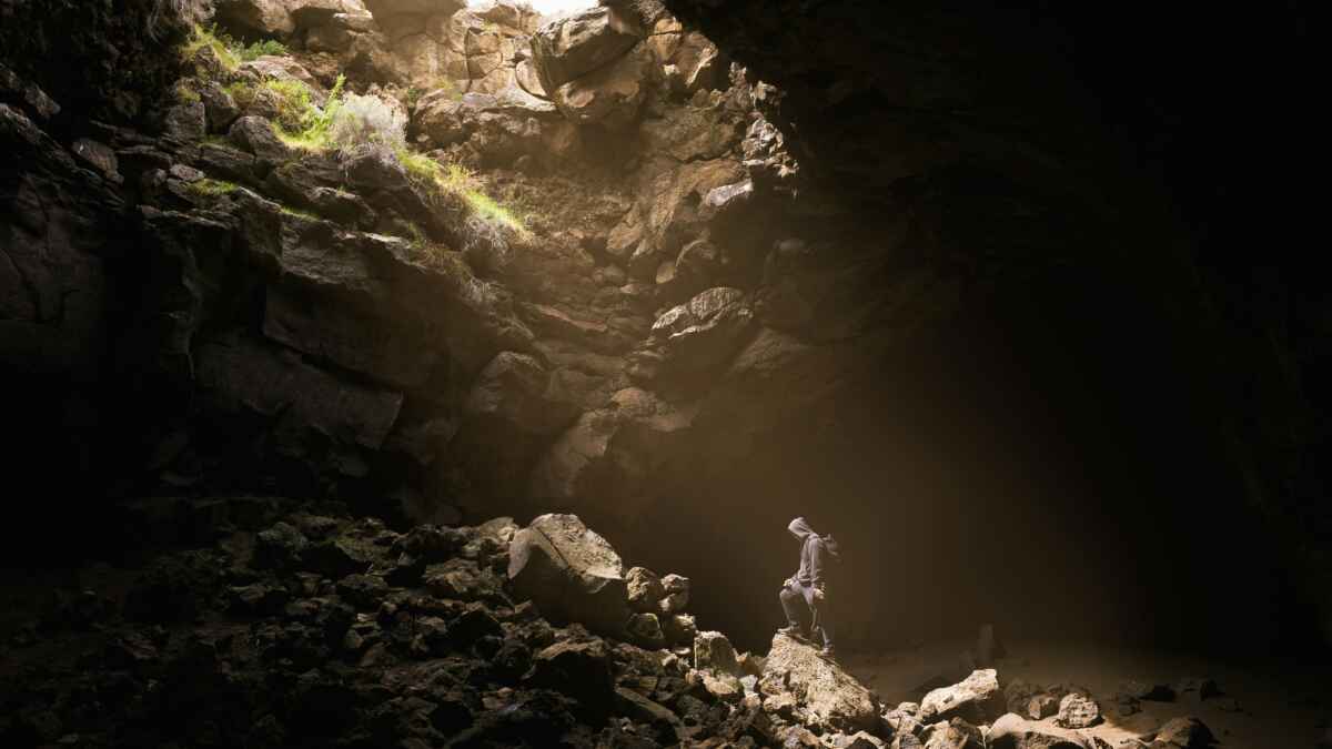 Person in Kapuzenpulli erkundet felsige Höhle mit Lichteinfall von oben, umgeben von dunklen Schatten und Felsschutt.