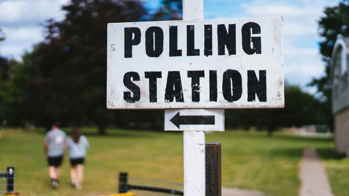 Schild mit der Aufschrift Polling Station zeigt nach links, im Hintergrund spazieren zwei Personen in einem Park.